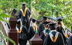 Graduates in full regalia walking across the Crim Dell bridge on a path through the woods