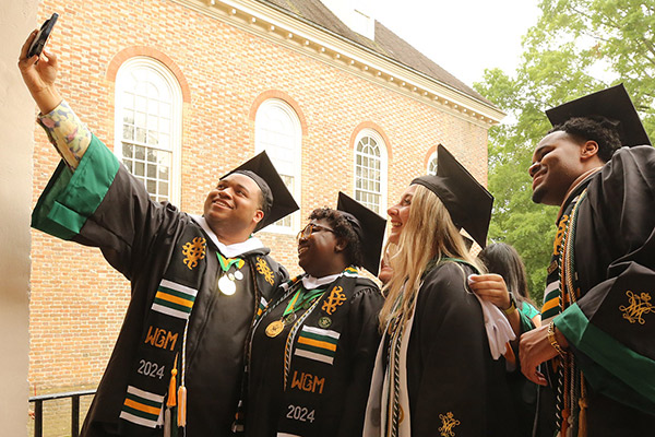 Student in full commencement attire taking a selfie with several other students arm in arm on the Wren Portico